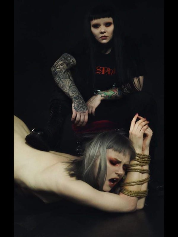 Two women in alternative fashion posing in a dramatic scene with one seated and the other bound on the floor.