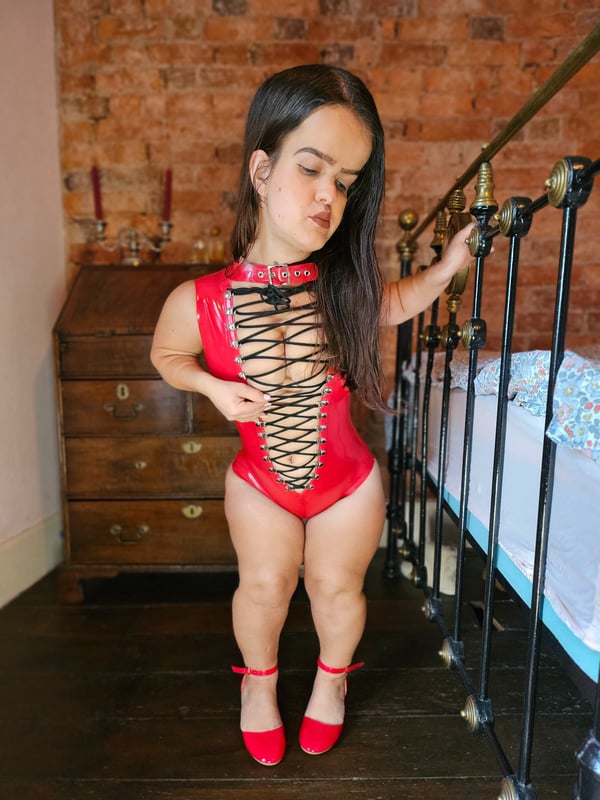 A petite woman standing in a vintage room with brick walls, wearing a red latex outfit and high heels, posing playfully near a bed.
