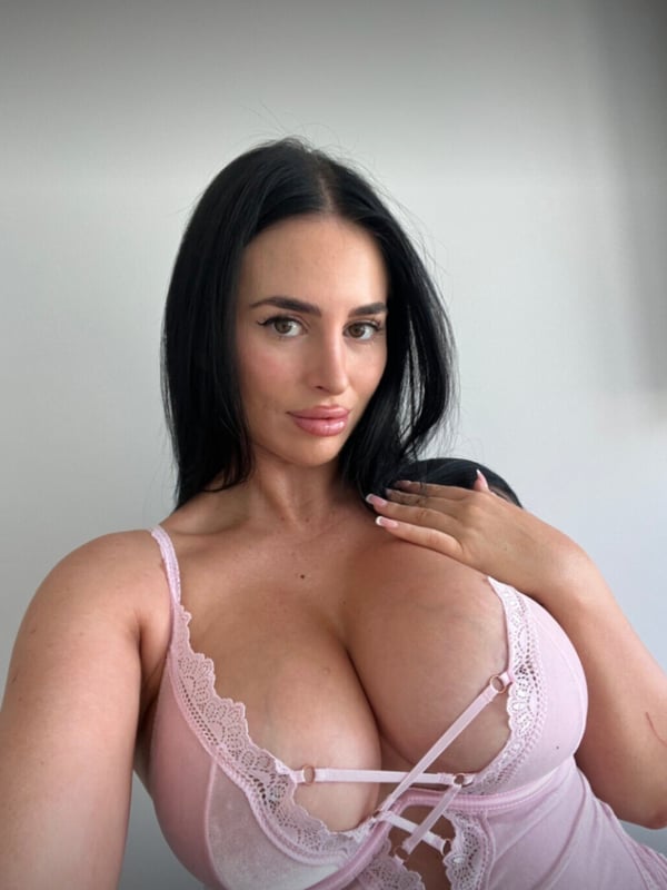 Close-up portrait of a woman with long dark hair wearing pink lace lingerie, smiling at the camera.