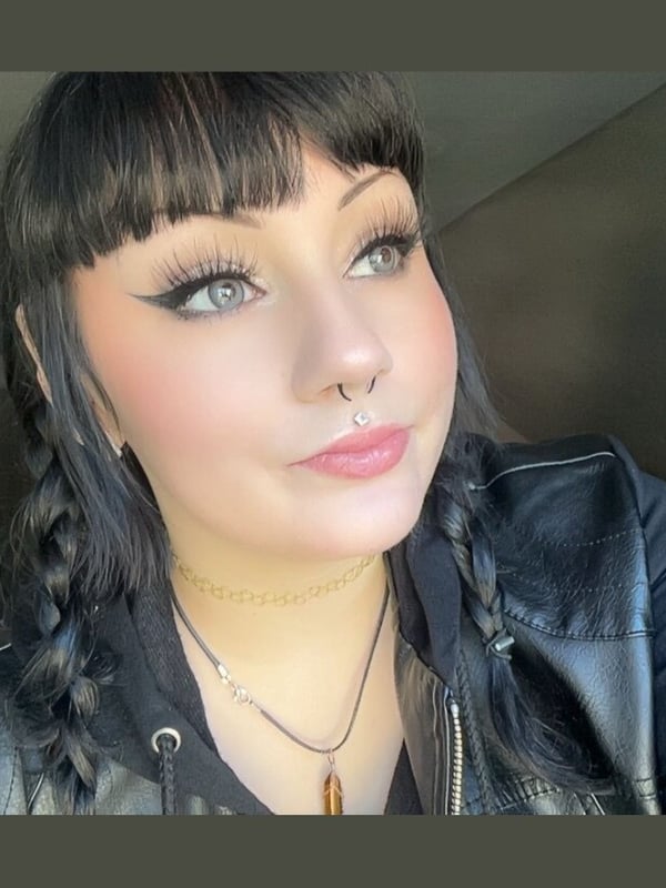 A young woman with dark braided hair, bangs, and facial piercings, wearing a black leather vest and gold necklace, posing indoors with a subtle smile.