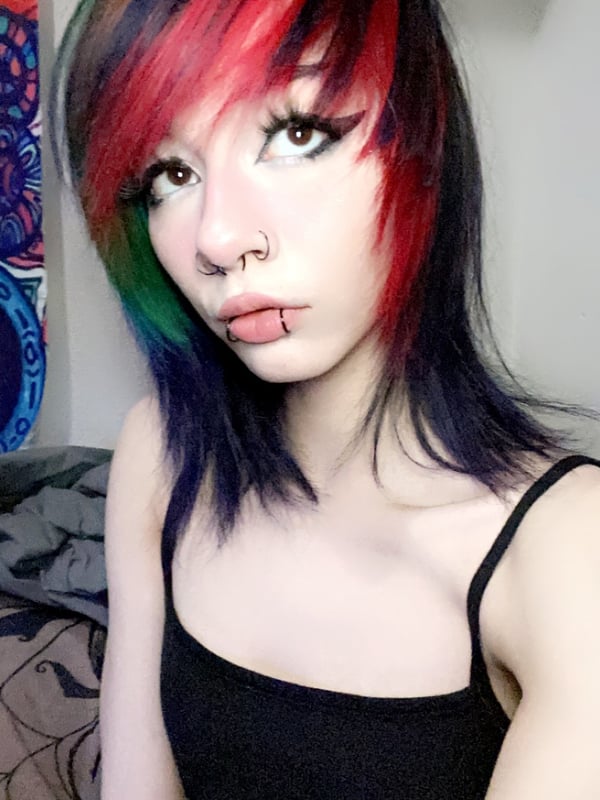 Young woman with pink hair and cat ears posing playfully near a crib in a colorful room with artistic wall decor.