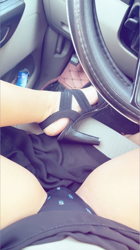Close-up view inside a car showing legs in high-heeled sandals resting on the dashboard near the steering wheel.