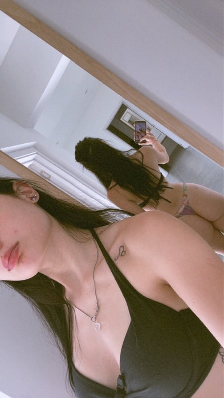 Young woman with long dark hair posing in a black bikini in front of a mirror in a bedroom.