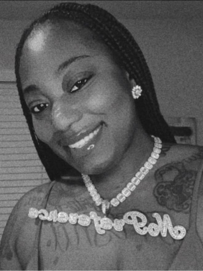 Black and white portrait of a smiling Black woman with braided hair, wearing pearl jewelry and a top with 'ROLLER YOUR ROLLS' text, posing indoors near a window.