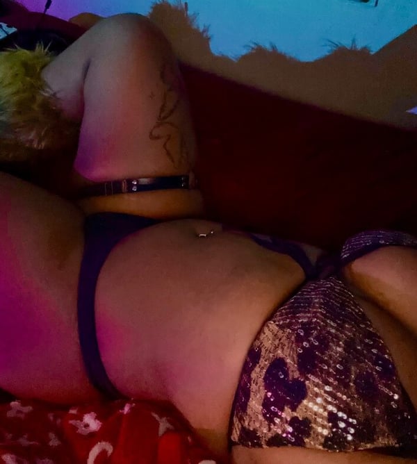 A person lounging on a bed in colorful swimsuit attire with tattoos and piercings visible, against a vibrant neon-lit background.
