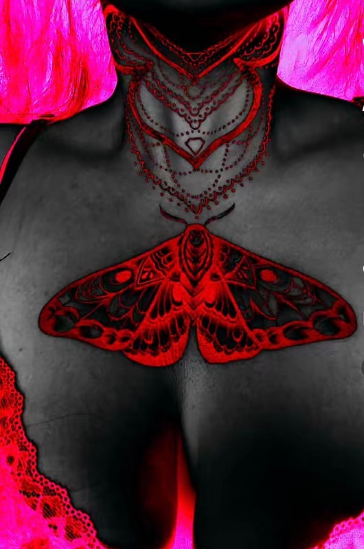 Close-up artistic portrait featuring intricate red jewelry around the neck and a large red butterfly tattoo on the chest, framed by pink lace elements.