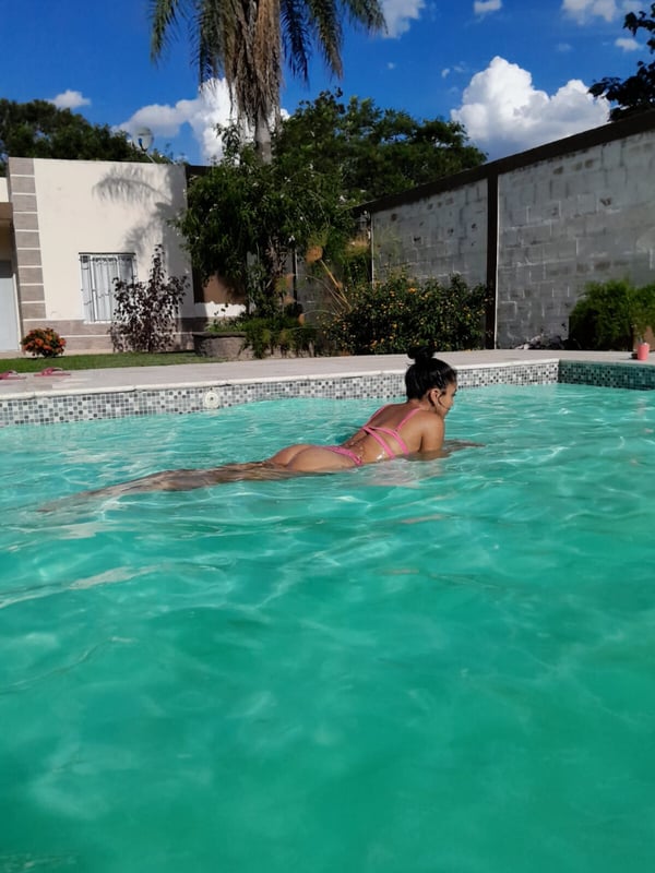 A woman lounging by a swimming pool in a pink swimsuit under a palm tree with a clear blue sky.