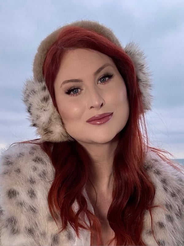 A person with long red hair wearing a fur-trimmed hat and coat, posing outdoors near a body of water.