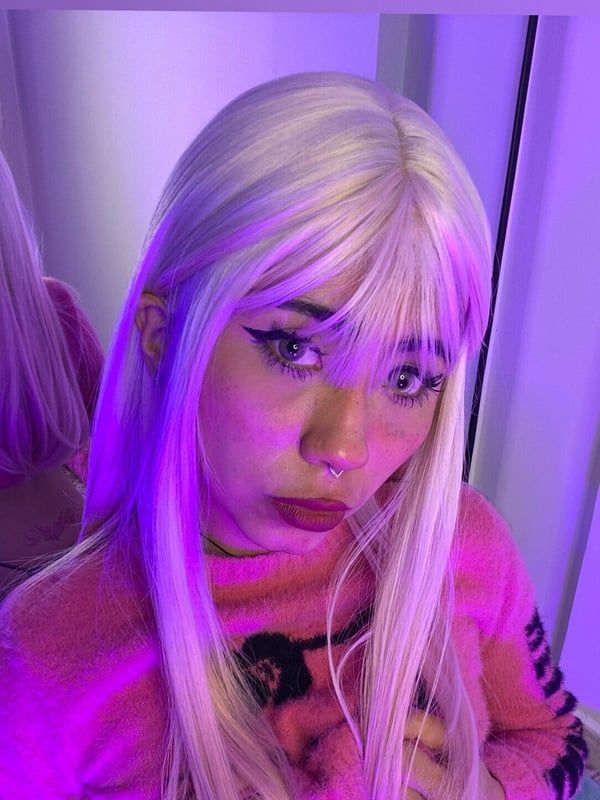 A close-up portrait of a person with long platinum hair and bangs wearing a pink fuzzy sweater under purple lighting.