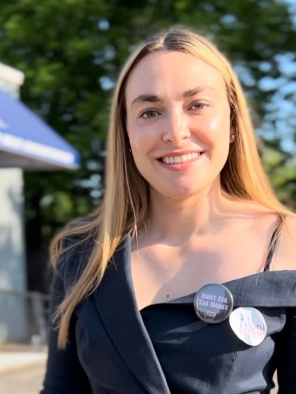 A woman with blonde hair smiling outdoors in front of a building with a blue awning, wearing a dark off-shoulder top and pins on her shoulder, with trees in the background.