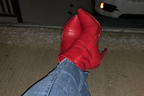 Close-up view of legs wearing tight jeans and red high-heeled boots positioned near a white car at night.