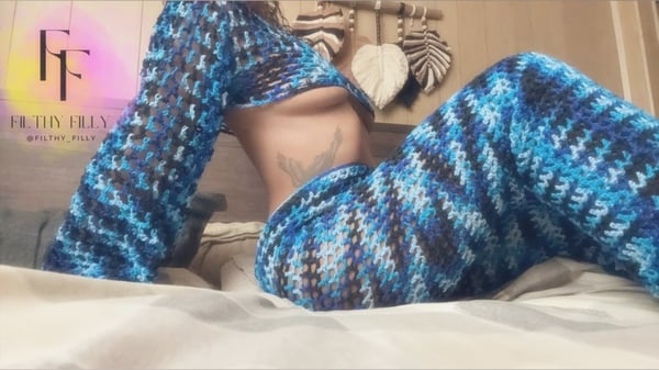 Woman posing on a bed in a colorful knitted outfit with decorative wall hangings in the background.