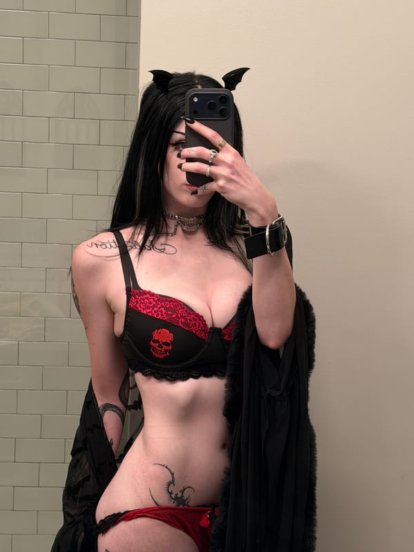 A person with long dark hair and glasses taking a mirror selfie in a cozy room with anime posters and decorations on the wall.