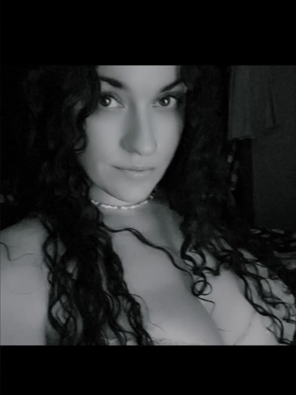 Black and white close-up portrait of a woman with long curly hair, wearing a necklace, gazing at the camera.