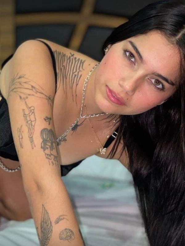 Close-up of a young woman with long dark hair and striking makeup, leaning forward on a bed in a black outfit with chains and jewelry.