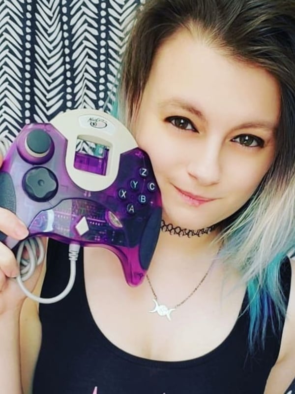 Young woman with colorful hair smiling while holding a purple gaming controller, wearing a black tank top and choker necklace against a dark striped backdrop.