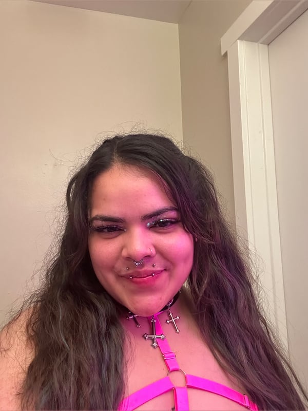 Young woman with long wavy hair, piercings, and choker necklace smiling in a room with pink walls.
