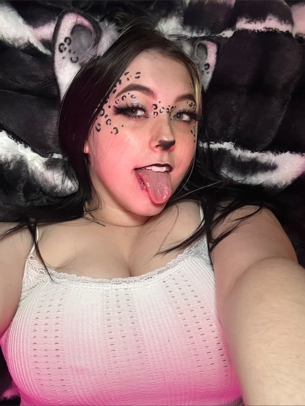 Young woman with cat ears, face paint, and dark hair sticking out her tongue in a selfie, dressed in a white lace top.