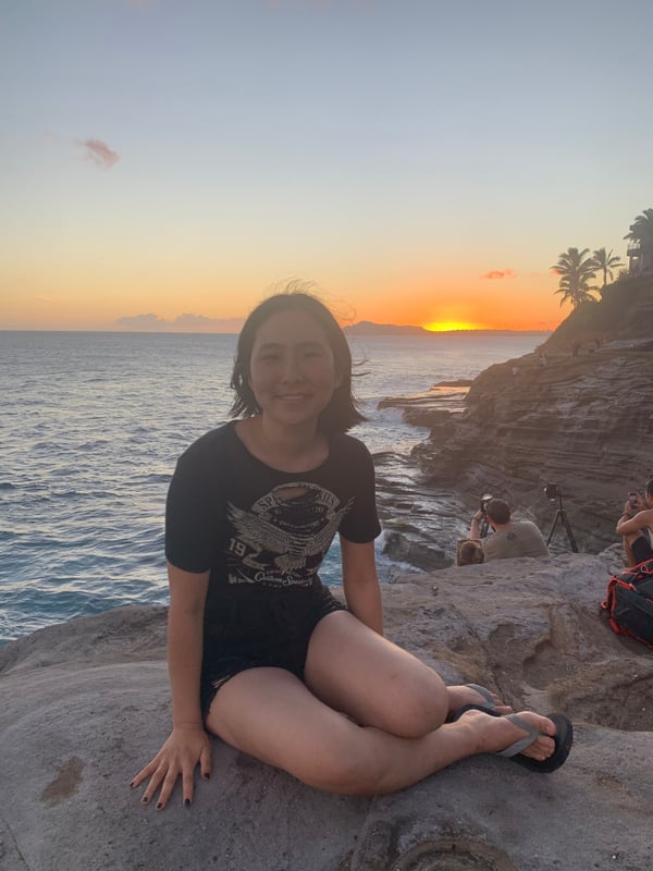 Young woman with short dark hair smiling while sitting on rocks at sunset overlooking the ocean and cliffs with palm trees.