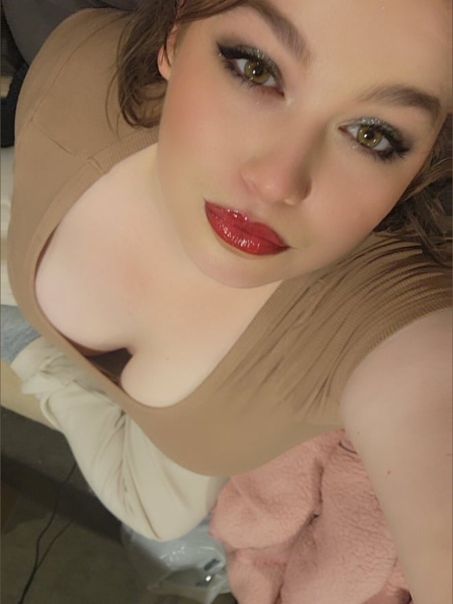 Close-up selfie of a young woman with brunette hair, green eyes, and red lipstick, wearing a low-cut beige top, looking up at the camera.