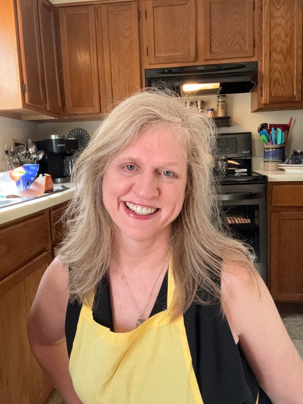 A woman with blonde hair smiling in a kitchen, wearing a yellow apron over a black top while standing near wooden cabinets and appliances.