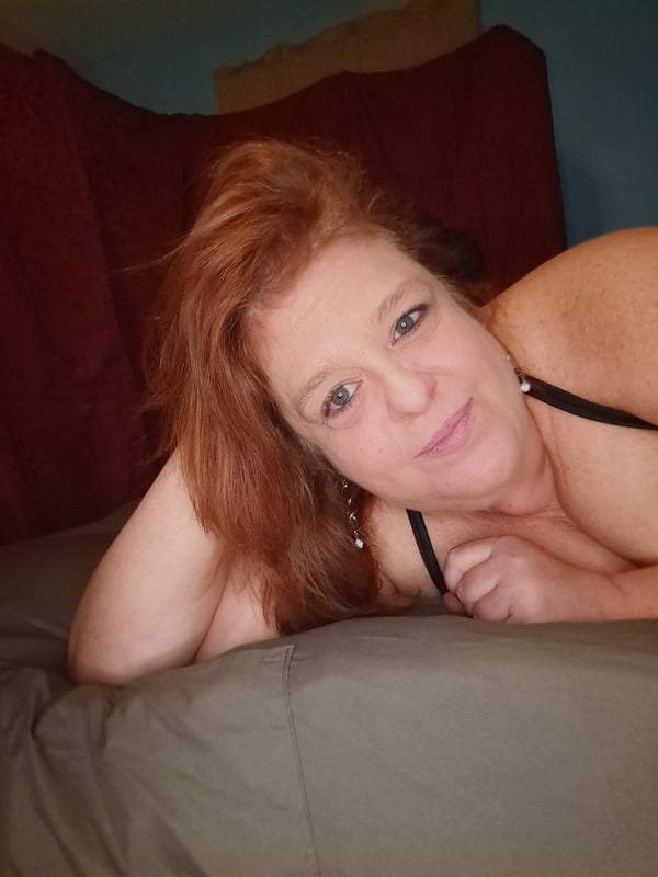 A woman with red hair smiling while leaning on a bed in a cozy bedroom setting.