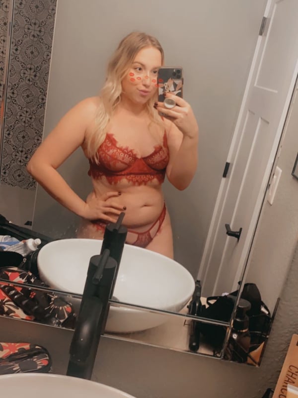 A woman with blonde hair posing in a red lingerie set in front of a bathroom mirror, holding a phone for a selfie.
