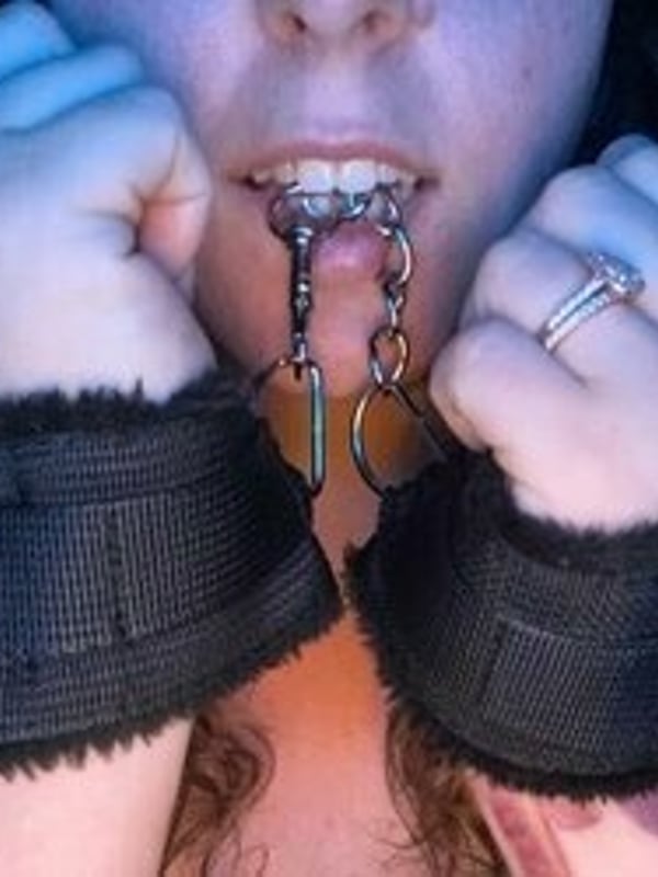 Close-up of a woman with curly brown hair holding metal chains near her mouth, wearing dark fuzzy wrist accessories.