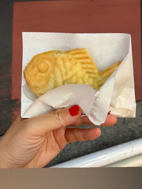 A hand holding a fish-shaped pastry wrapped in white paper on a red surface.