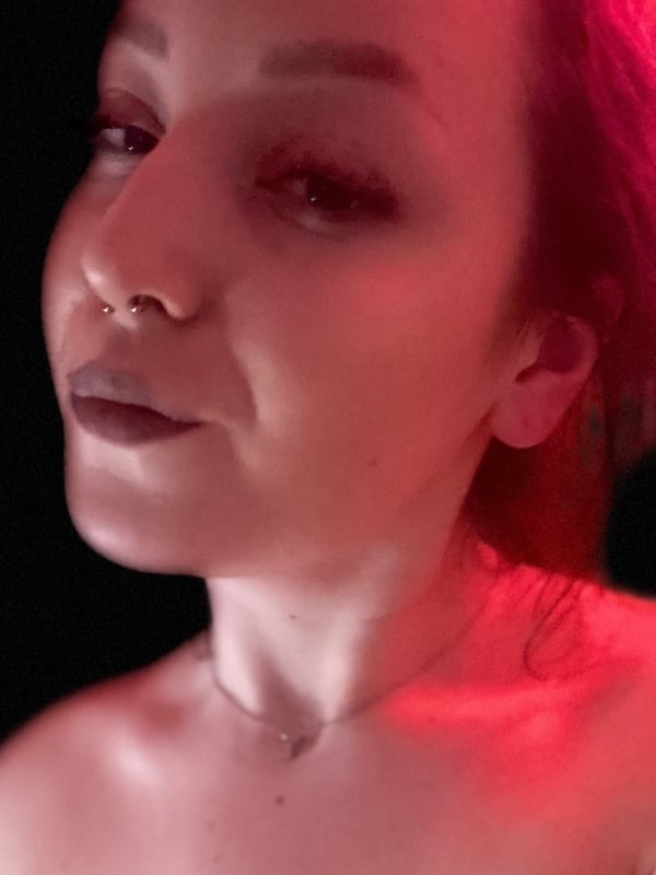 Close-up portrait of a young woman with red hair, piercing, and subtle makeup, illuminated by red lighting against a dark background.