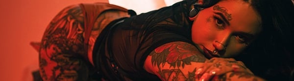 A woman with tattoos poses seductively on a red background, wearing form-fitting clothing.