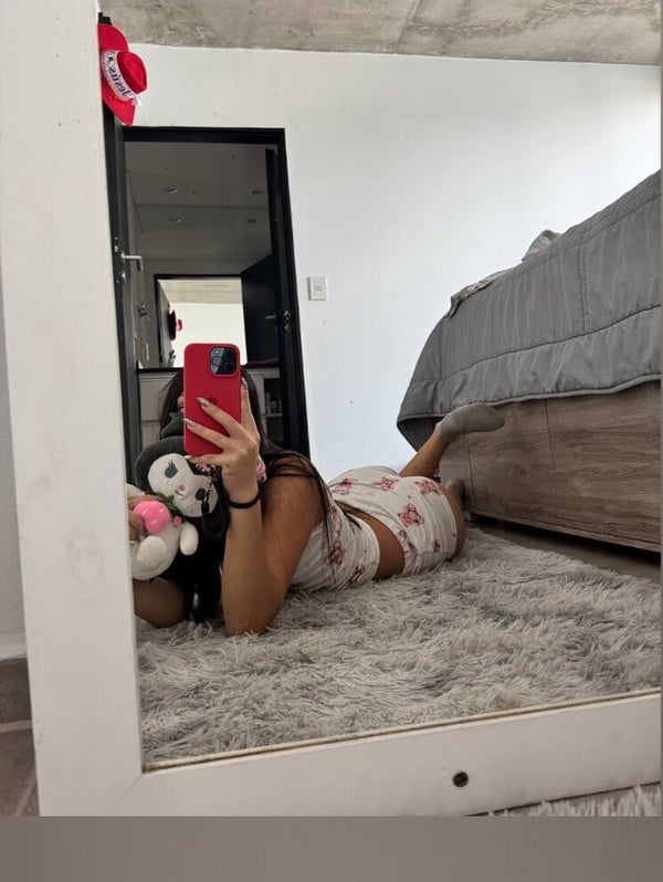 Young woman lying on the floor in front of a mirror, wearing a floral-patterned outfit and red hat, holding a phone and stuffed animal.