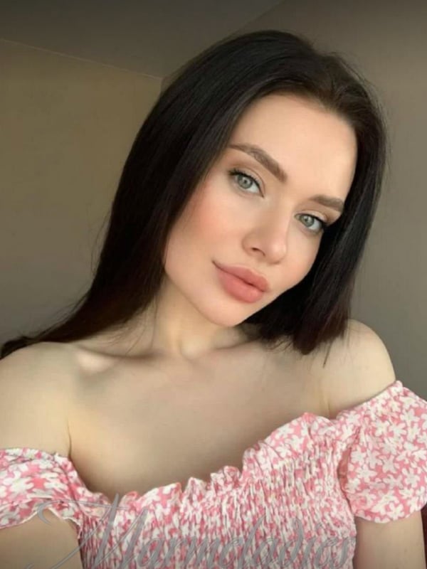 Close-up portrait of a young woman with long dark hair, green eyes, and subtle makeup, wearing an off-shoulder pink top.