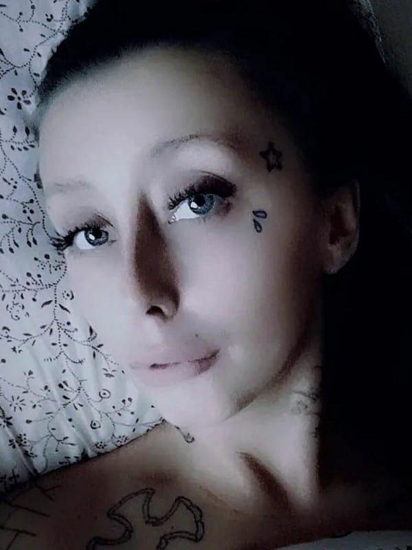 Close-up portrait of a woman with dark hair, facial tattoos, and expressive eyes, lying on a bed with floral bedding.