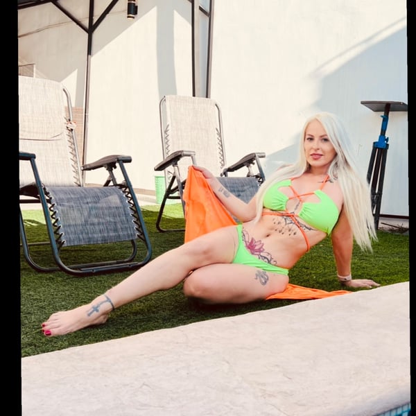 Woman in a green bikini sitting by the poolside on green grass, with lounge chairs and an outdoor patio in the background.