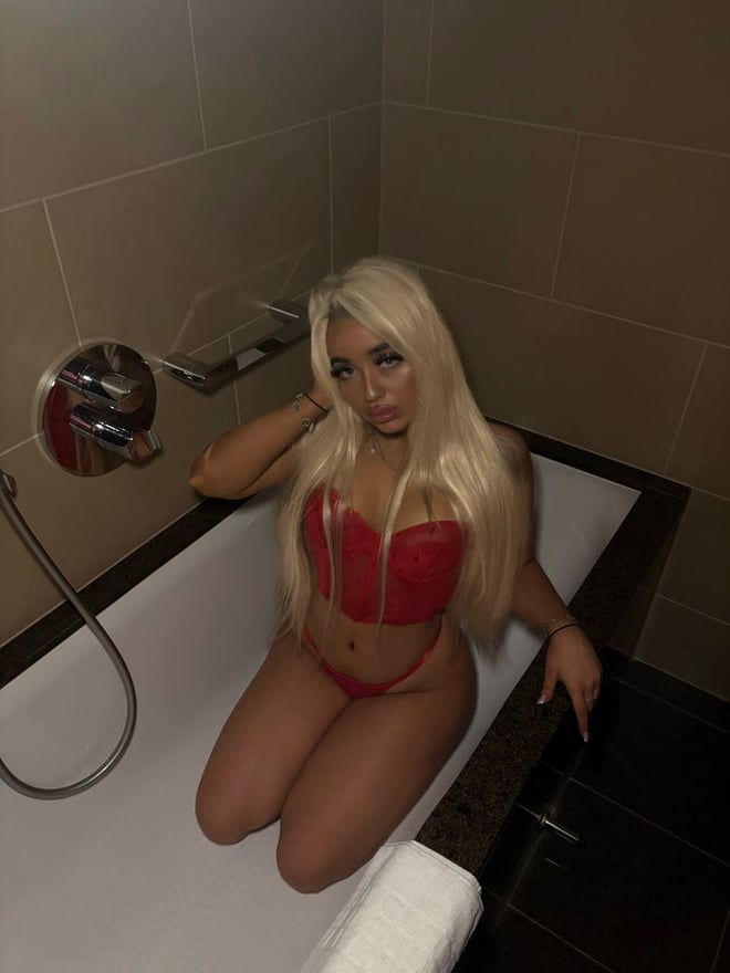 A blonde woman posing in a red outfit while seated in a bathtub.