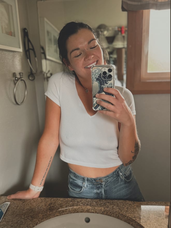 A smiling woman taking a mirror selfie in a bathroom, wearing a white crop top and jeans.
