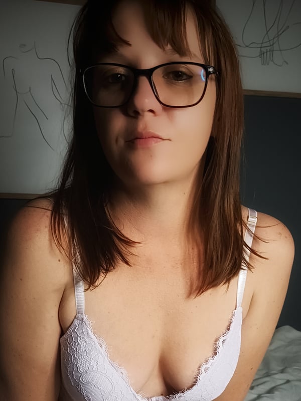 A woman with glasses and long brown hair, wearing a white lace top, posing indoors against a wall.