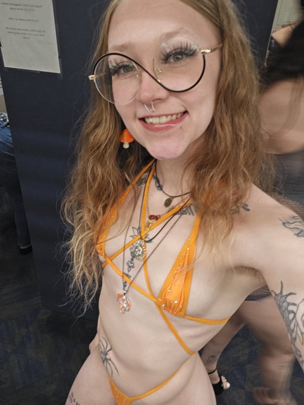 A smiling woman with freckles, blonde wavy hair, and glasses stands in a bright orange bikini top and necklace against a dark booth background.