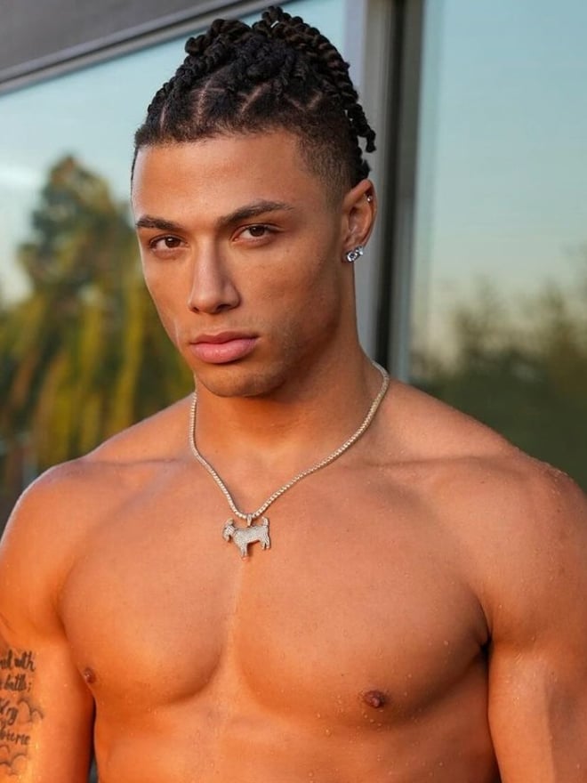 A muscular person with braided hair and jewelry poses in front of a window, with a tattoo visible on the upper arm.