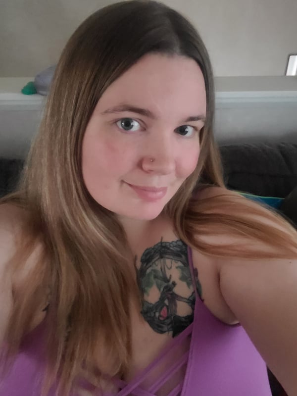 A woman with long brown hair smiling at the camera, wearing a purple top, with a prominent tattoo visible on her chest, posing indoors.