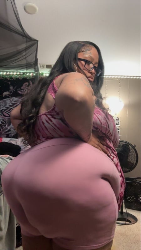 A woman with long dark hair wearing glasses and a pink top and shorts poses confidently from behind in a bedroom with sheer curtains and soft lighting.