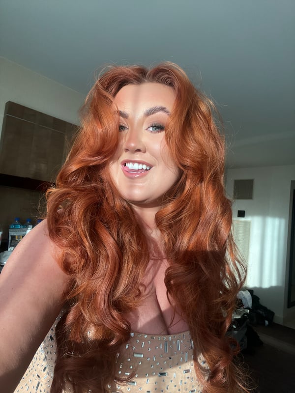 A woman with long wavy red hair smiling at the camera in a sparkly top, posing indoors with a casual background.