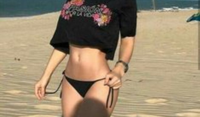 A woman posing confidently on a beach in a cropped floral top and bikini bottoms, with palm trees and ocean in the background.