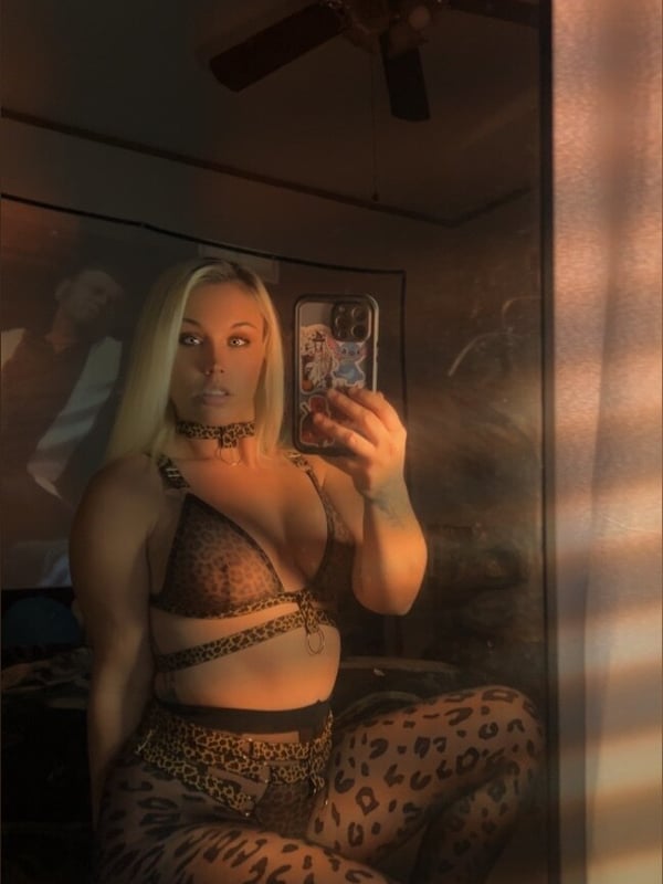 A woman with long blonde hair taking a mirror selfie in a bedroom, wearing a leopard print outfit and choker necklace.