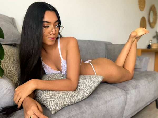 A woman with long dark hair and glasses posing on a gray couch in a light-colored room.