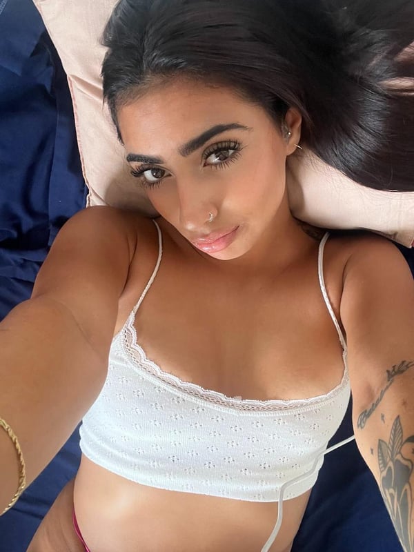 Young woman with long dark hair lying on a bed, wearing a delicate white lace top, smiling at the camera.