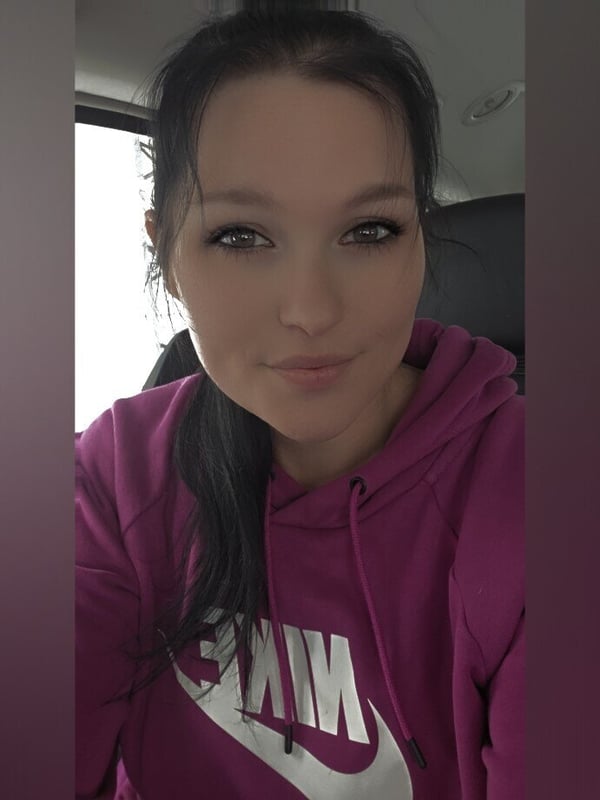A young woman with dark ponytail smiling in a car, wearing a pink Nike hoodie.
