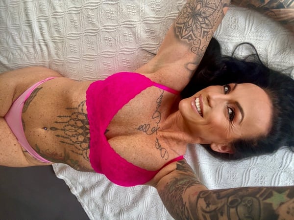 A woman with long dark hair and tattoos poses smiling on a bed, wearing pink lingerie and extending her arms upward.