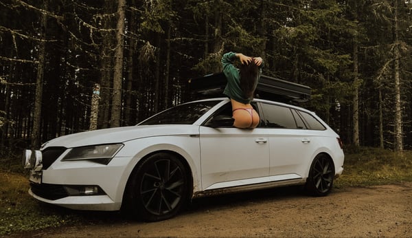 A woman in green clothing leaning out of the open door of a white station wagon parked in a forested area.